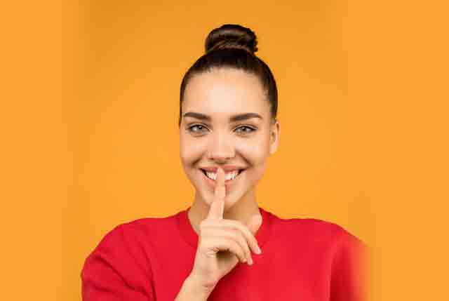 A photo of a smiling Latina woman in a red sweater with her hair in a bun, holding up her index finger to her lips